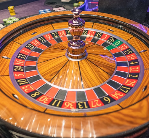 A roulette wheel with alternating red and black numbered pockets on a wooden platform. The wheel contains a small white ball resting in one of the pockets, indicating gameplay. Surrounding the wheel are stacks of colorful poker chips, suggesting a casino environment.