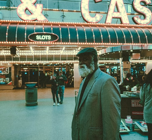 An older man with a gray beard and wearing a suit and hat stands in front of a brightly lit casino. The background features a large illuminated sign reading 'CASINO' and 'SLOTS' among other words, with numerous lights outlining the entrance. Several other people are visible in the background, engaged in various activities, some walking, some standing, and one looking at something on display.